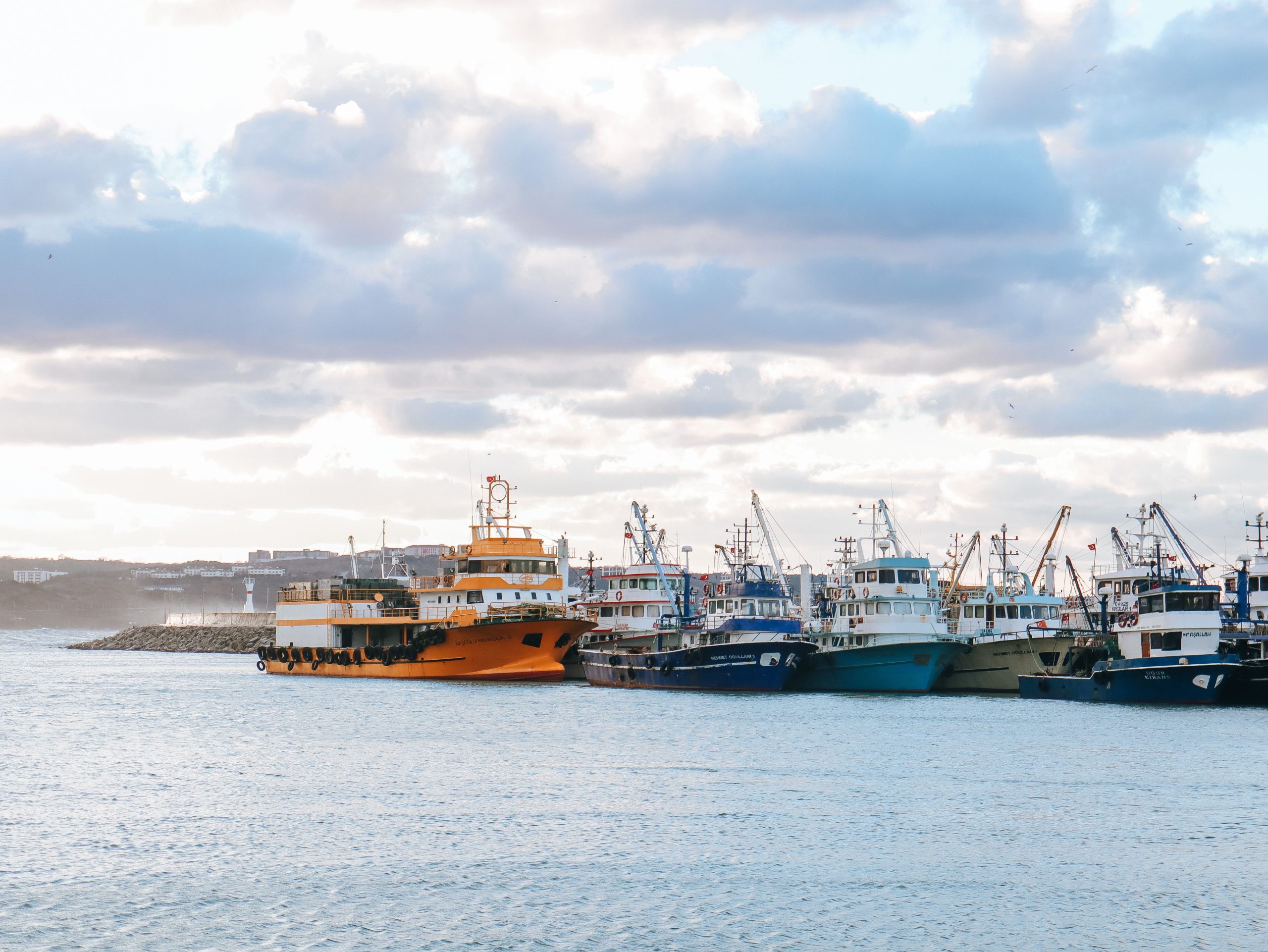 A variety of colorful fishing boats docked at a harbor during daytime under partly cloudy skies.